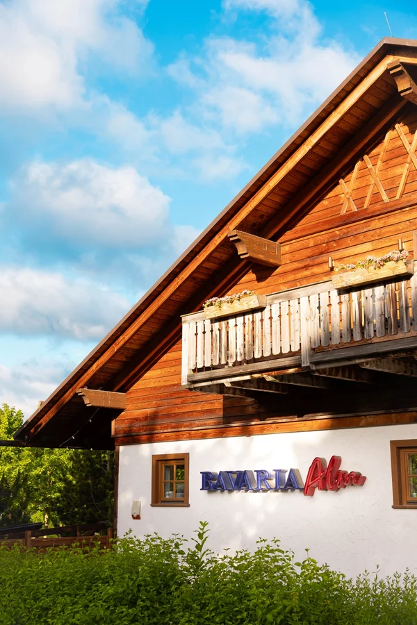 Außenansicht der Bavaria Alm mit rustikaler Holzfassade und Balkon unter blauem Himmel.
